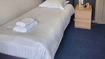 A neatly made bed with white linens and towels, a bedside table with a bottle and remote, and a wall-mounted light fixture.