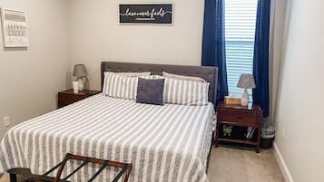 A bedroom with a bed, a nightstand, a lamp, a chair, and a wall with a quote.