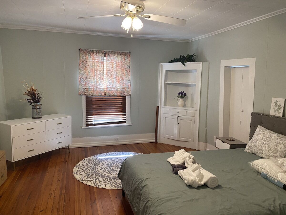 A bedroom with a bed, a white dresser, a window with blinds, a ceiling fan, and a built-in shelving unit.