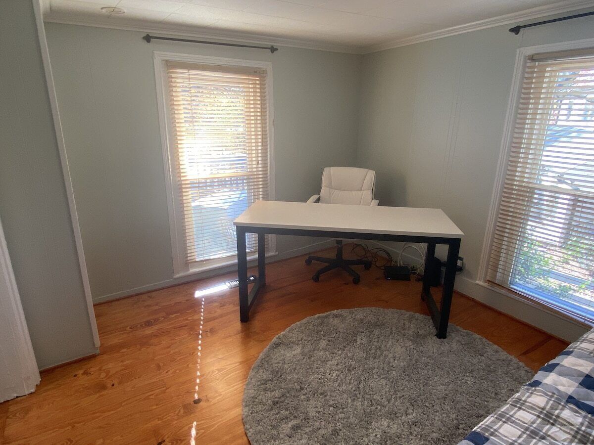 Une pièce avec un plancher en bois, un bureau blanc avec une chaise, un tapis gris et deux fenêtres équipées de stores.