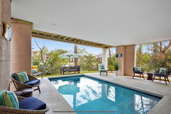 A covered outdoor swimming pool with lounge chairs and umbrellas, surrounded by greenery and a clear view of the sky.