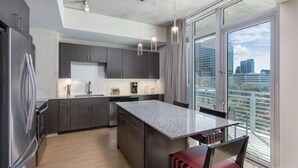 Une cuisine moderne avec des meubles de couleur sombre, un plan de travail en granit et un grand îlot central. On y trouve un réfrigérateur, un évier et une fenêtre offrant une vue sur la ville.