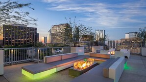 Un salon sur le toit, doté de mobilier moderne et d’un foyer central, offrant une vue panoramique sur la ville.