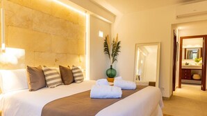 Una habitación de hotel con una cama grande, un cabecero, una mesita de noche, un espejo y un baño visible a través de una puerta abierta.