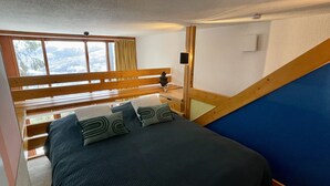 A bedroom with a bed, a wooden bench, a window with curtains, and a blue wall. - Apartment Mezzanine Perriand Arcs 1800 Valley View (Les Arcs 1800)