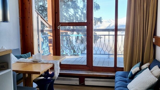 A cozy room with a wooden floor, a blue sofa, a wooden table, and a large window with a view of trees and mountains. - Apartment Mezzanine Perriand Arcs 1800 Valley View (Les Arcs 1800)