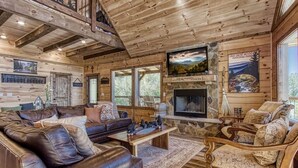 A log cabin interior with a stone fireplace, leather sofas, and a large TV. - Huge Mountain LOG Cabin with Amazing Views (Gatlinburg)
