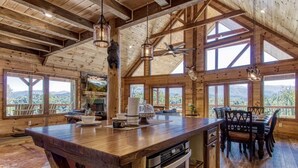 A spacious kitchen with a large island, wooden beams, and large windows offering a view of the outdoors. - Huge Mountain LOG Cabin with Amazing Views (Gatlinburg)