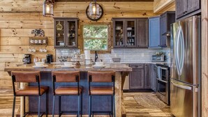 A rustic kitchen with wooden walls, a central island with bar stools, dark cabinets, and stainless steel appliances. - Huge Mountain LOG Cabin with Amazing Views (Gatlinburg)