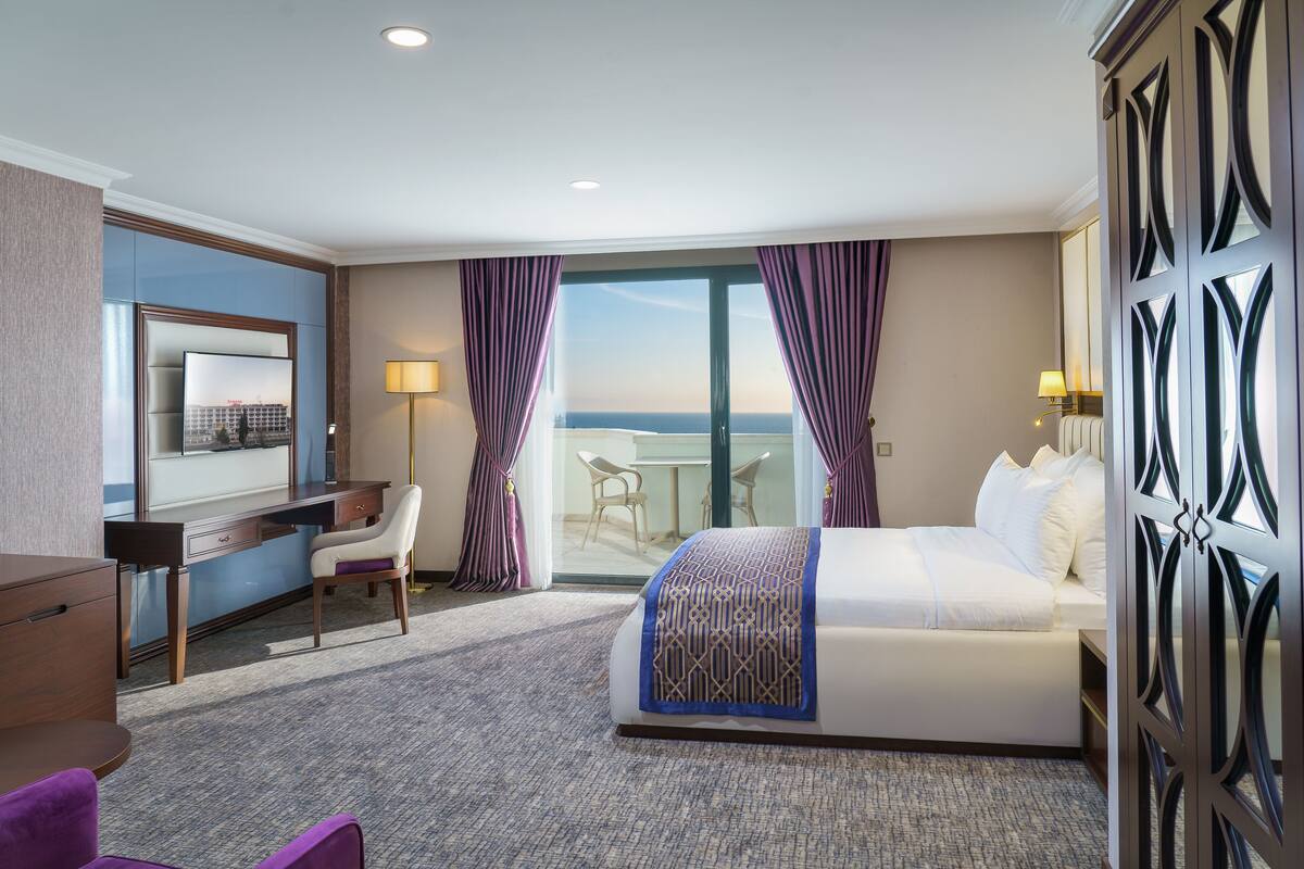 Een hotelkamer met een bed, een bureau, een stoel en uitzicht op zee door een glazen deur.