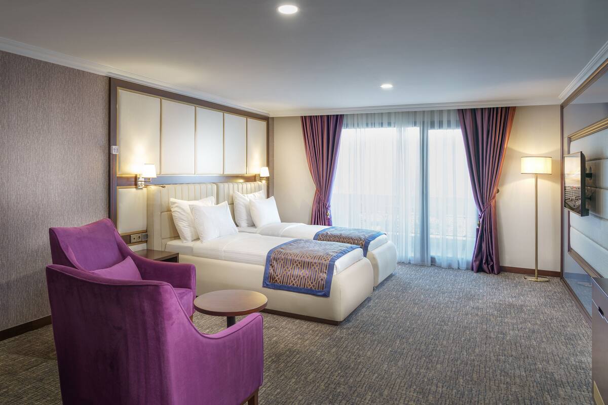 Een hotelkamer met een groot bed, twee nachtkastjes, een paarse fauteuil, een kleine ronde tafel en een grote spiegel.
