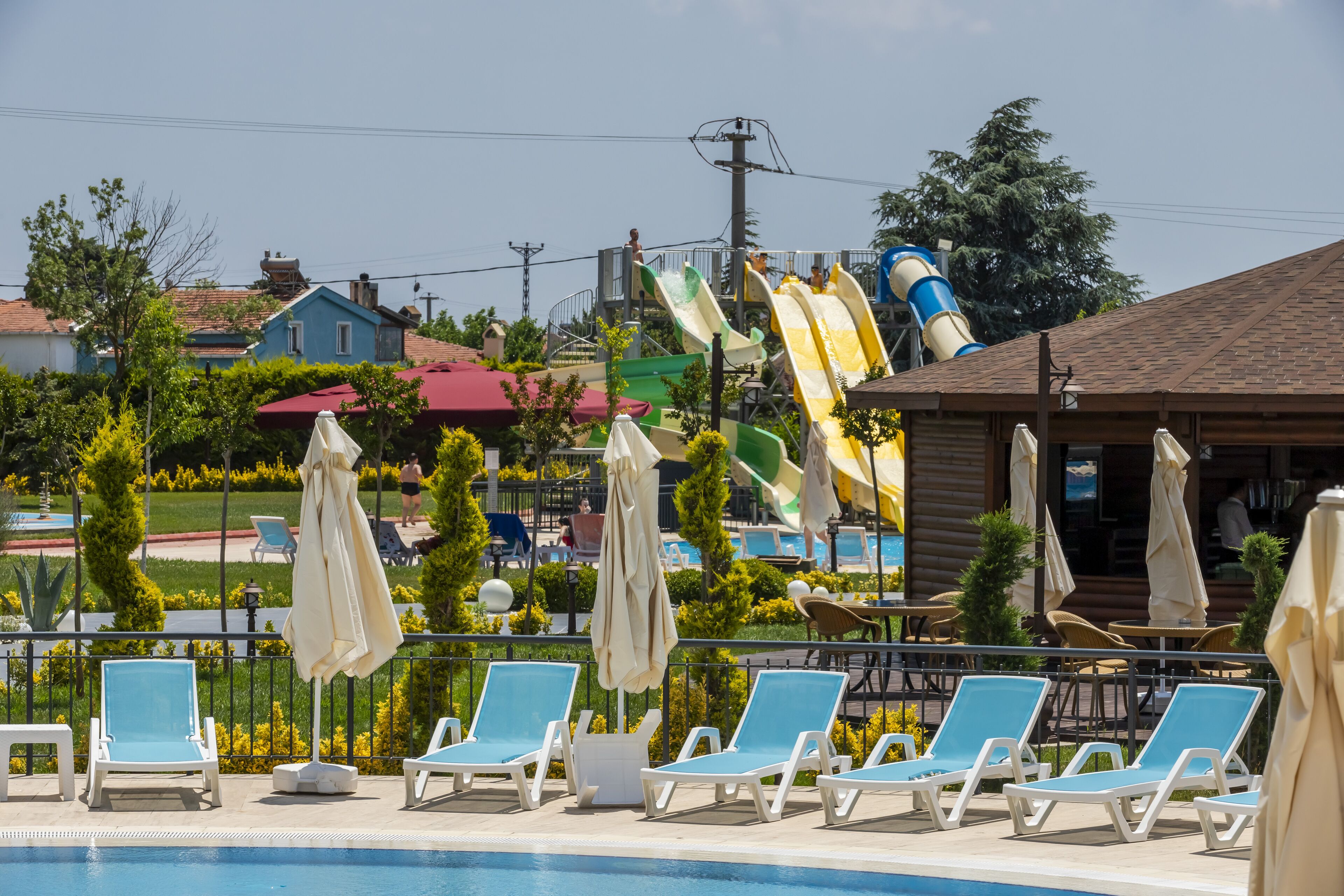 Un área de piscina con sillones de descanso y sombrillas, un tobogán de agua y un edificio con techo rojo.