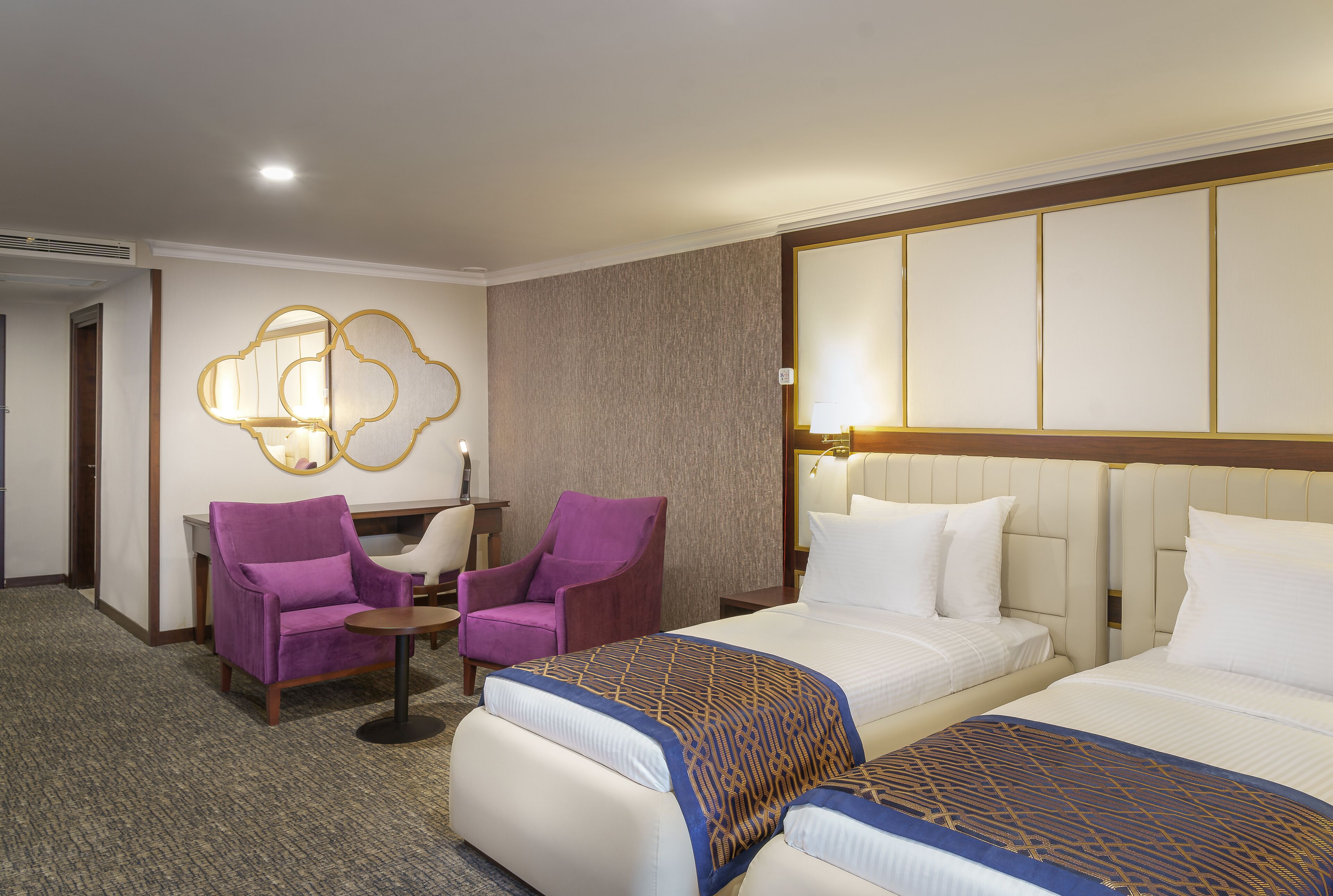A hotel room with two beds, a purple armchair, a small round table, and a wall-mounted lamp.