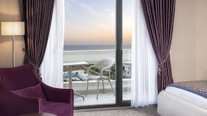 A hotel room with a balcony, a purple chair, a white bed, and a view of the sea.