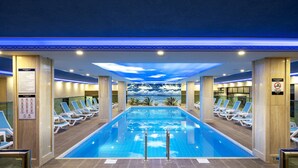 An indoor swimming pool with a view of a beach and palm trees, surrounded by lounge chairs and a ceiling with a cloud design.