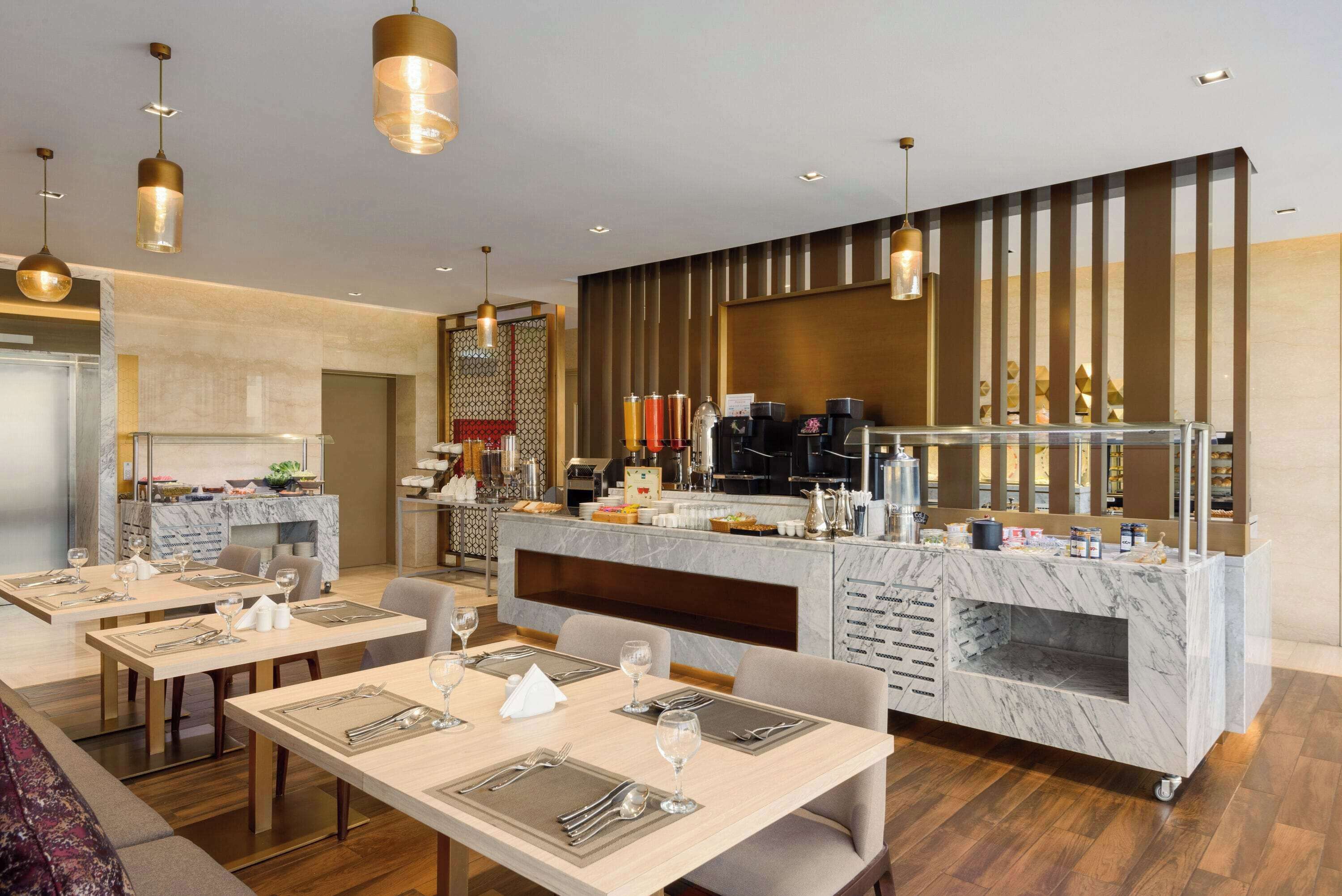 A modern hotel dining area with marble countertops, multiple dining tables set with silverware, and a central bar area with a marble island.