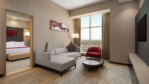 A hotel room with a bed, a grey sofa, a red chair, a round coffee table, and a large window with a view. - Ramada By Wyndham Riyadh King Fahd Road (Riyadh)