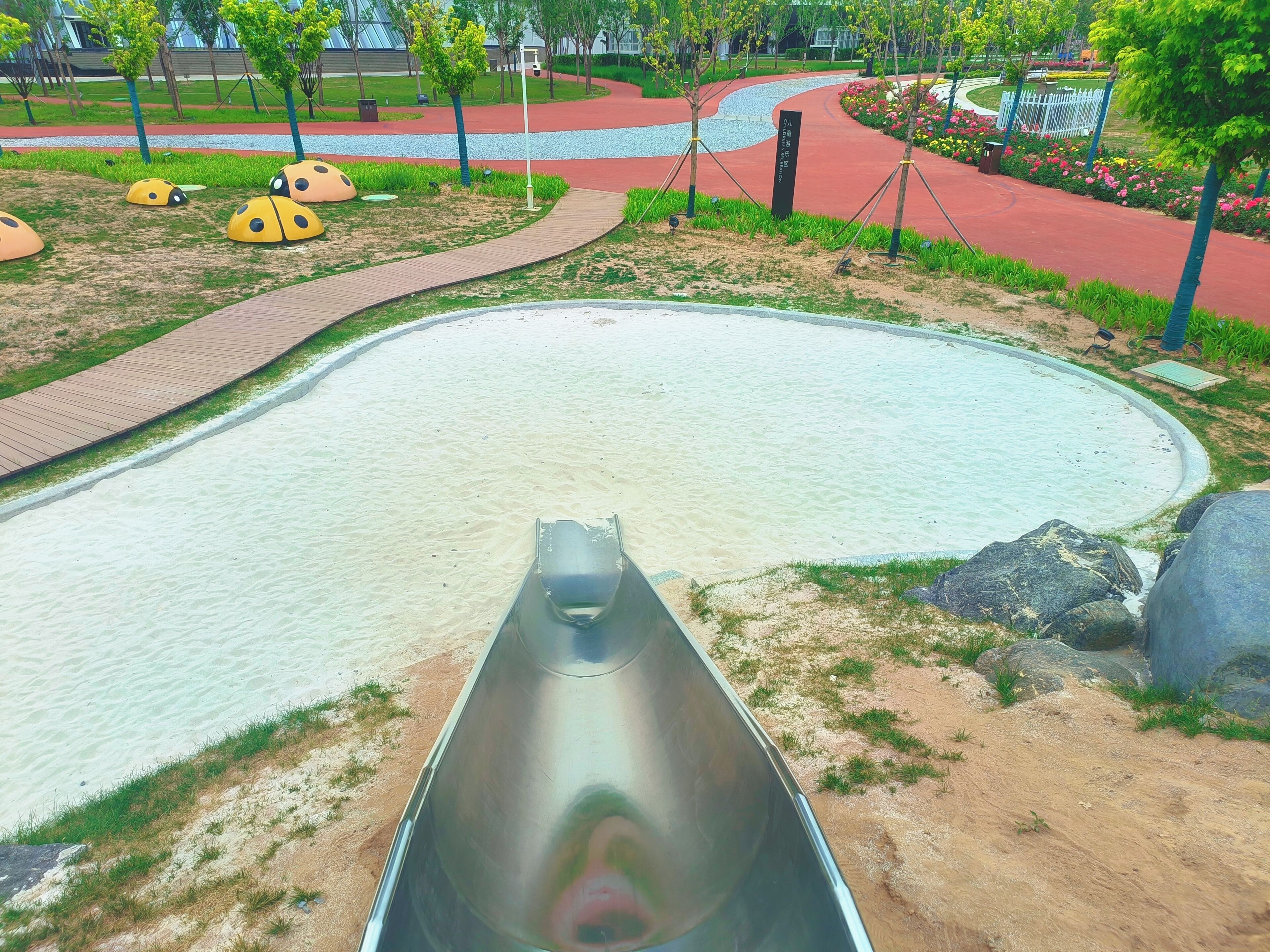 Un espace de jeux avec un toboggan, un chemin sinueux et un espace aménagé avec des arbres et des fleurs.