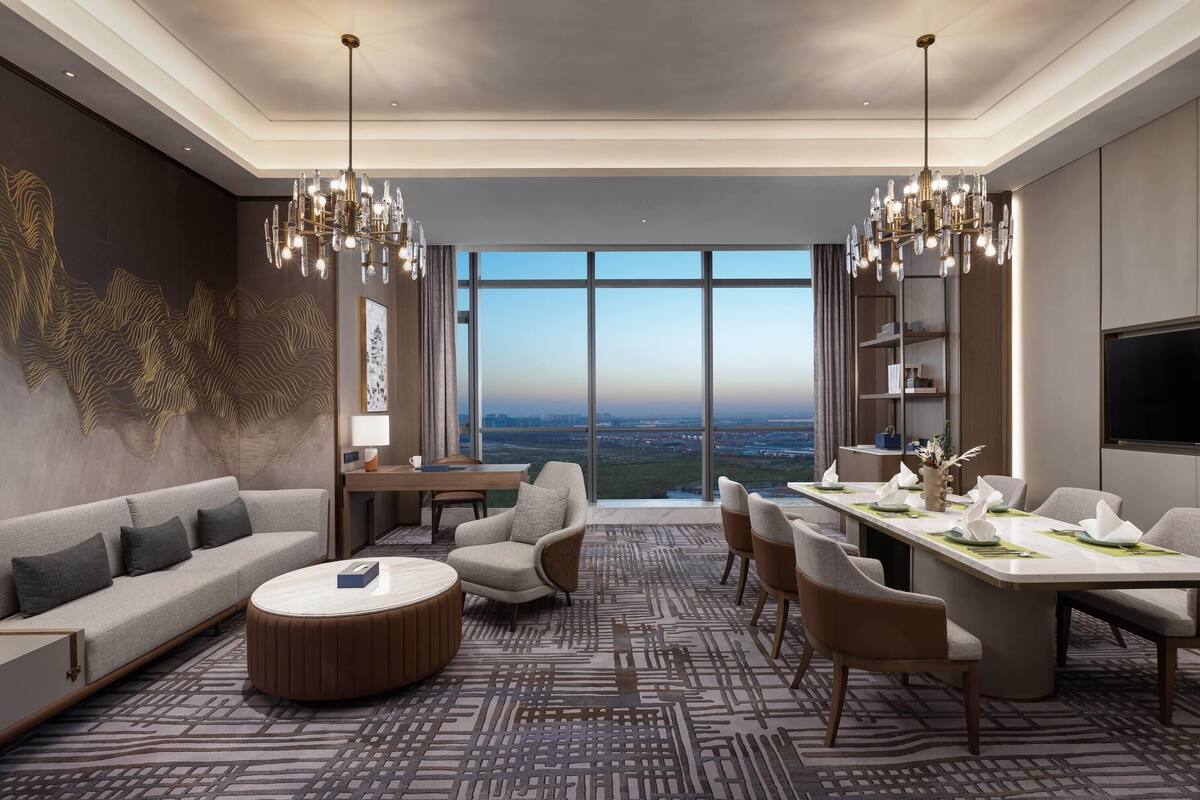 Una habitación de hotel moderna con un ventanal grande, un sofá, una mesa de comedor y una araña.