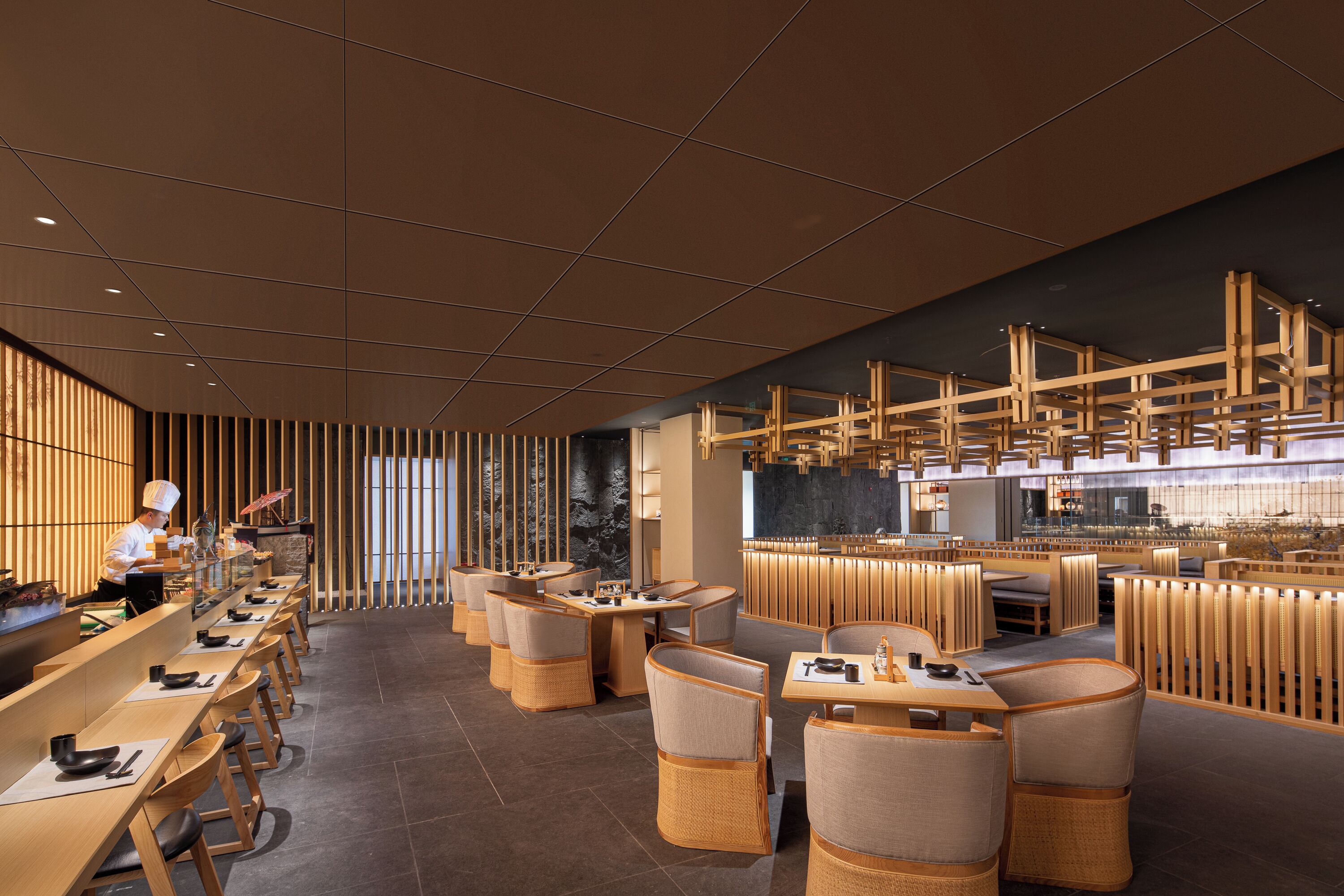 Un restaurante moderno con largas mesas comunitarias y cabinas individuales, un chef preparando comida y un techo alto con vigas de madera.