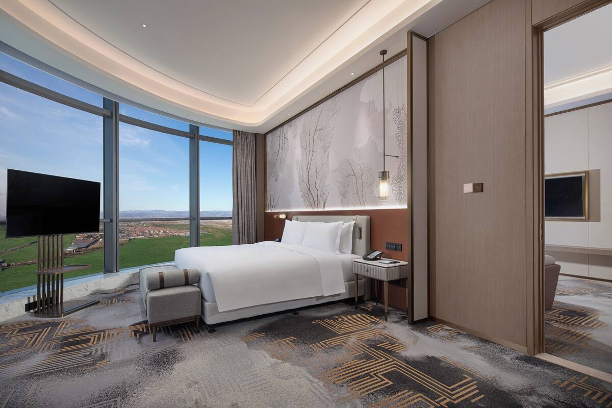 Habitación de hotel moderna con una cama grande, televisor de pantalla plana, mesita de noche con lámpara y vistas a un paisaje urbano a través de amplios ventanales.