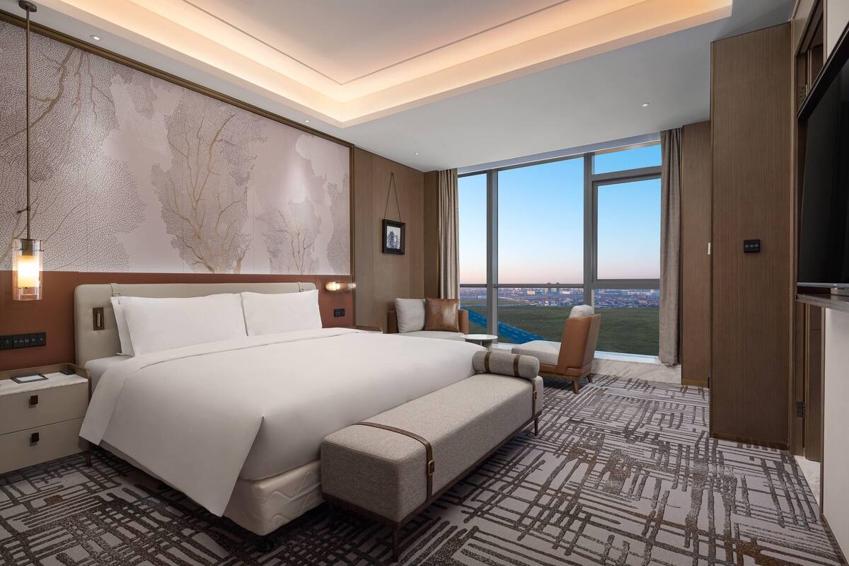 Una habitación de hotel moderna con una cama grande, un banco, un escritorio y vistas a la ciudad.