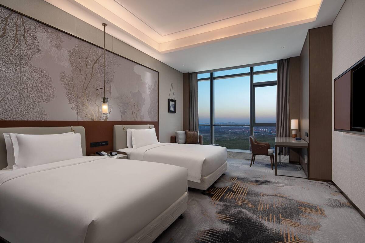 Habitación de hotel con dos camas, un escritorio, una silla, un amplio ventanal con vista a la ciudad y una obra de arte colgada en la pared.