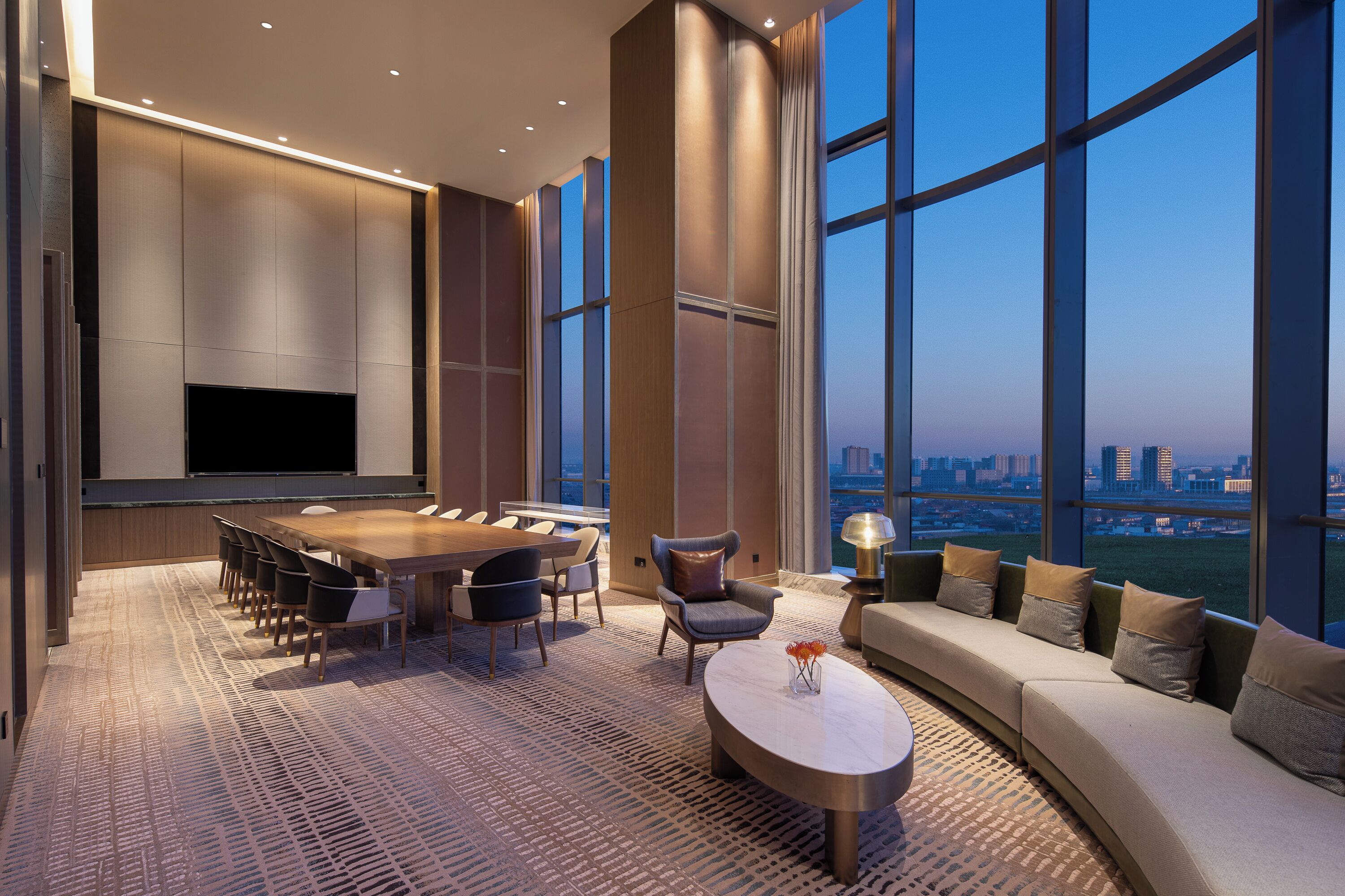 A modern hotel room with a large dining table, comfortable seating, a flat-screen TV, and a sofa with a view of the cityscape.
