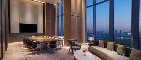 A modern hotel room with a large dining table, comfortable seating, a flat-screen TV, and a sofa with a view of the cityscape.