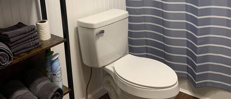A bathroom with a white toilet, a striped shower curtain, a framed picture on the wall, and a shelf with towels and toiletries.