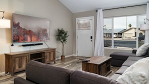 Un salon doté d’un téléviseur à écran plat, d’un meuble de rangement en bois, d’un canapé gris et d’une grande fenêtre offrant une vue sur une maison et une plage.