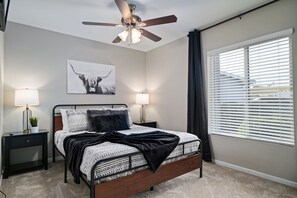 Une chambre à coucher comprenant un lit, deux tables de chevet, un ventilateur de plafond, une fenêtre avec des stores et une œuvre d’art accrochée au mur.