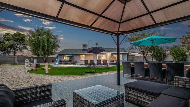 Une terrasse extérieure couverte, aménagée avec des meubles en osier et une table en verre, donnant sur un jardin arrière paysager avec une maison en arrière-plan.