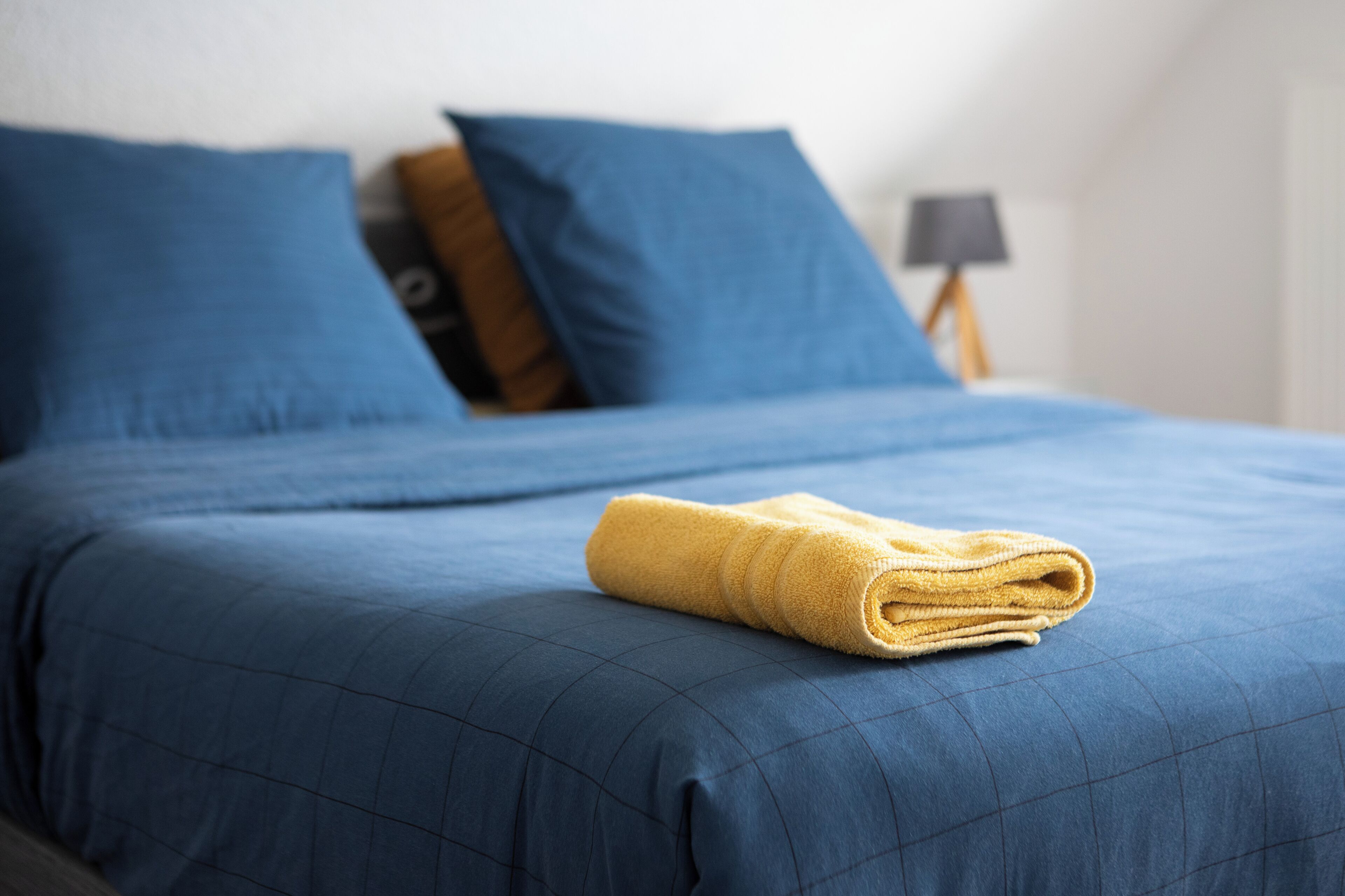 A neatly made bed with blue bedding and a rolled yellow towel on top.