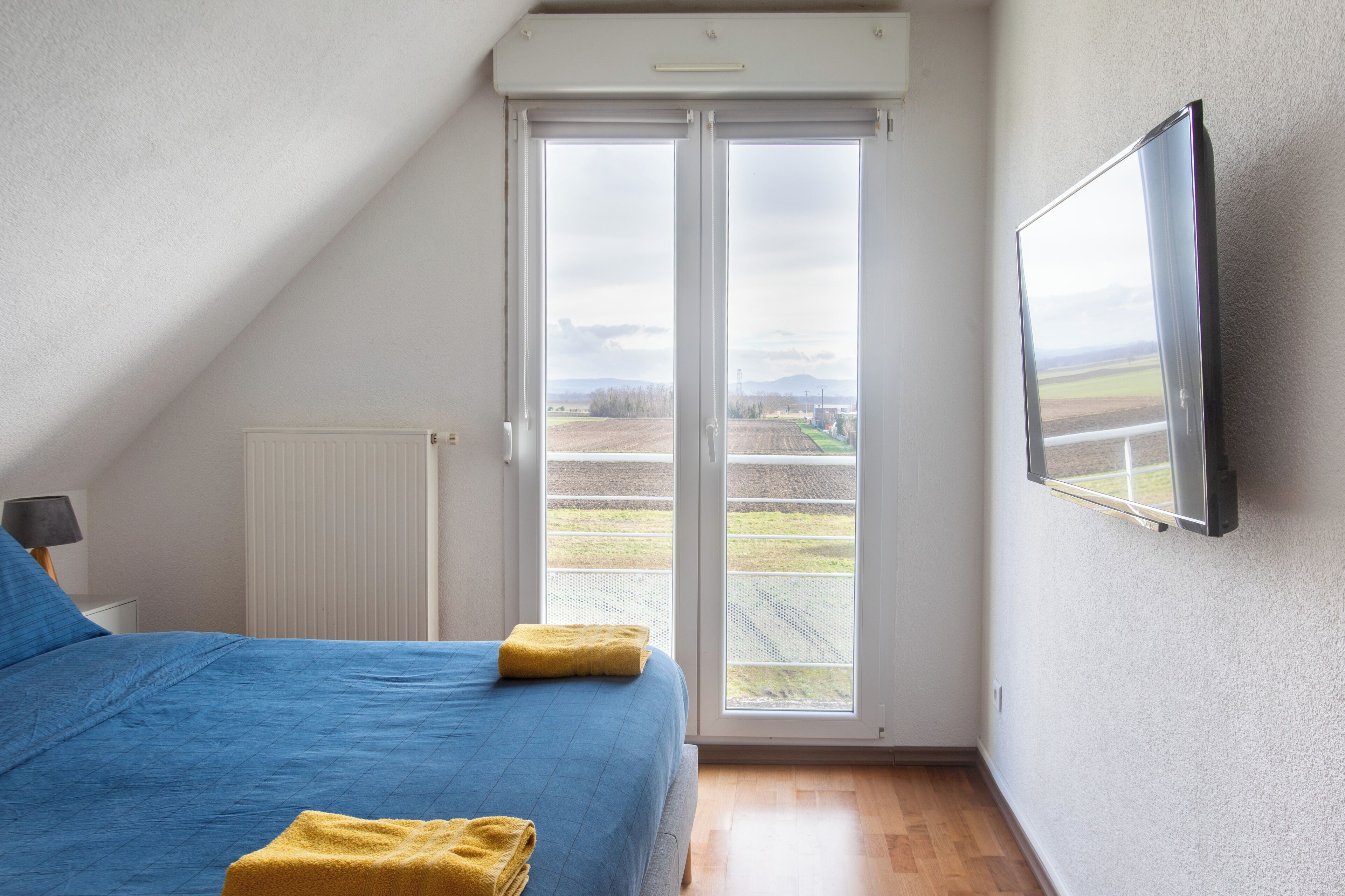 A bedroom with a large bed, a window with a view of a field, and a mounted TV on the wall.