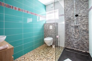 Een badkamer met blauwgroene tegels, een wit toilet, een douche met glazen wanden en een houten wastafelmeubel.