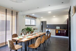 Een moderne eetruimte met een lange tafel voor zes personen, omringd door zes stoelen. Achtergrond toont een keuken met een wasmachine en een magnetron.