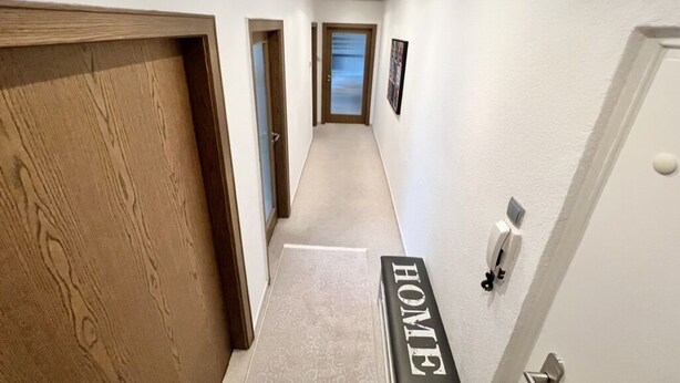 Een gang met een houten deur links, een witte muur met een 'HOME' bord, en een deur rechts.