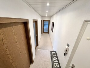 Un couloir avec une porte en bois sur la gauche, un mur blanc sur lequel est inscrit le mot « MAISON », et une porte sur la droite.