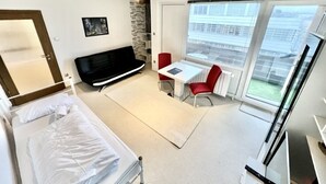 Een moderne hotelkamer met een bed, een bureau, een rode stoel en grote ramen met uitzicht op de stad.