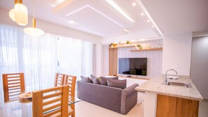 A modern living room with a grey sofa, wooden chairs, a glass dining table, a kitchen area with a sink, and a TV mounted on the wall. - 14th Floor Modern Ocean, Beach & Pool View Apartment in Juan Dolio (Guayacanes)