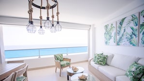 A balcony with a view of the sea, a white sofa with green pillows, a wooden dining table, and a chair with a green cushion. - 14th Floor Modern Ocean, Beach & Pool View Apartment in Juan Dolio (Guayacanes)
