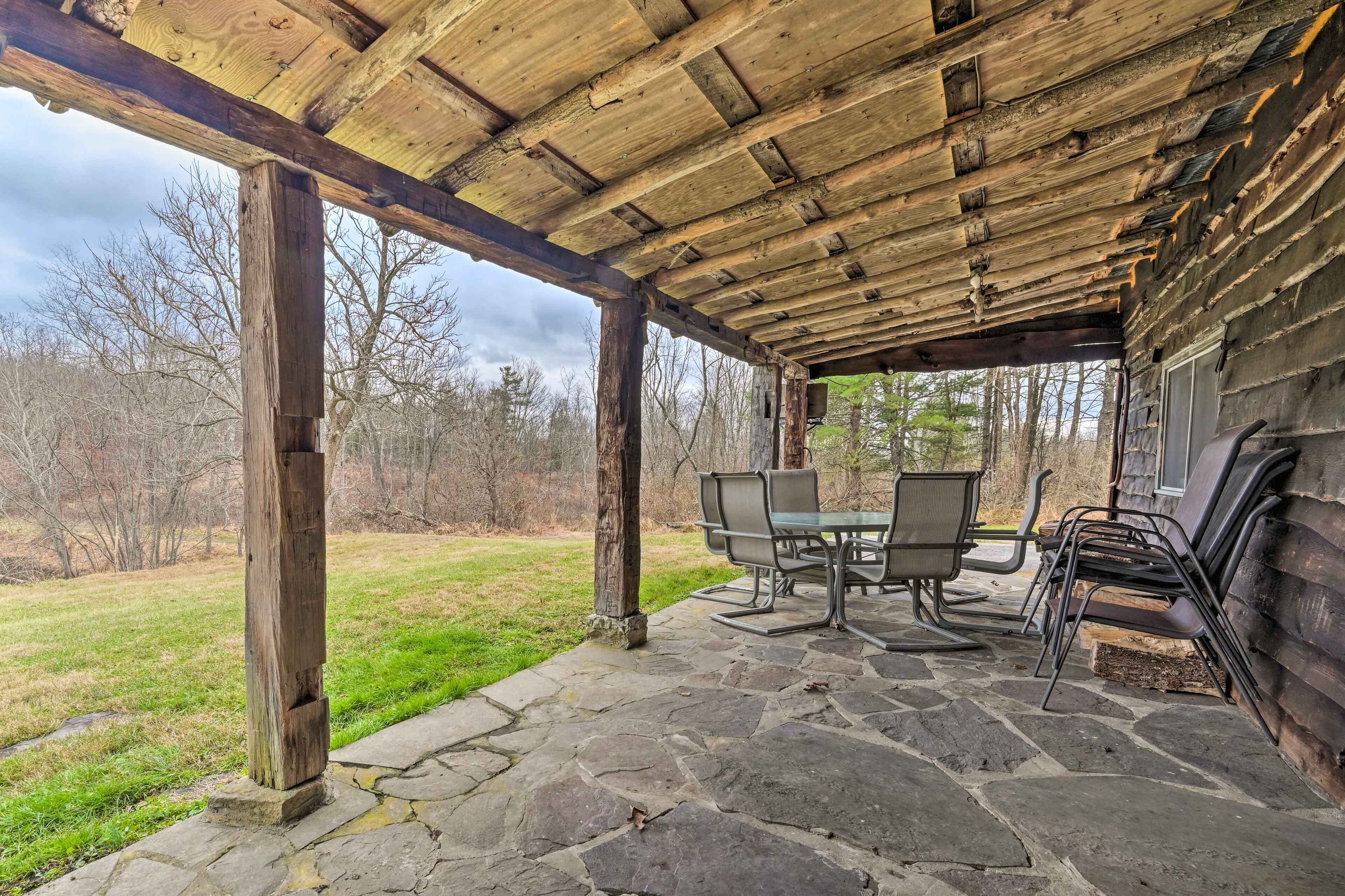 A covered outdoor seating area with stone flooring and wooden beams, overlooking a grassy landscape.