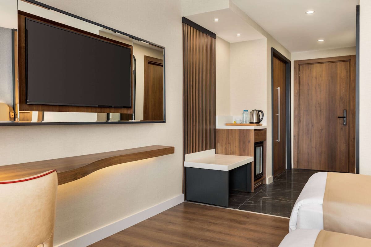 A modern hotel room with a flat-screen TV mounted on the wall, a wooden reception desk, a small sink, and a door leading to another room.