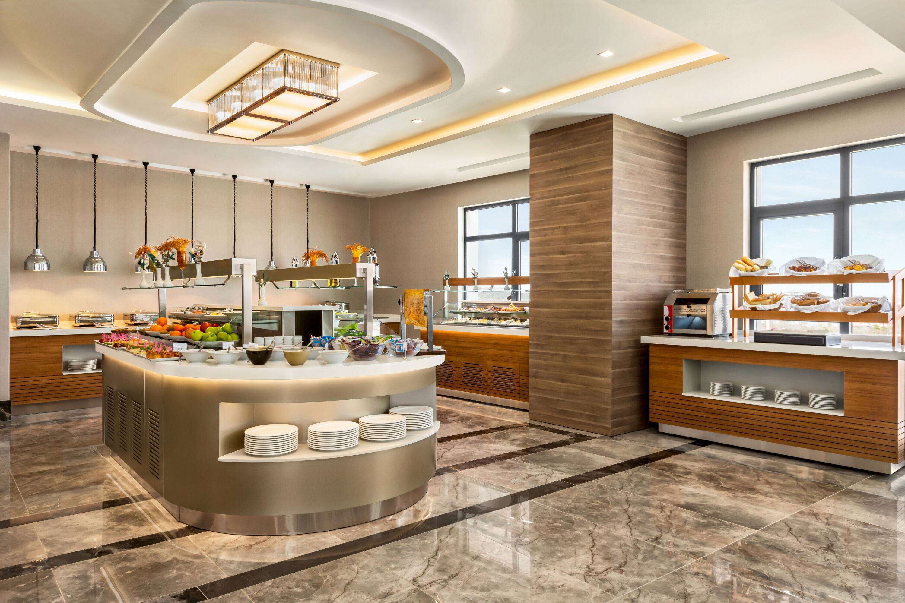 Cocina de hotel moderna con un bufé central, suelo de mármol e iluminación colgante.