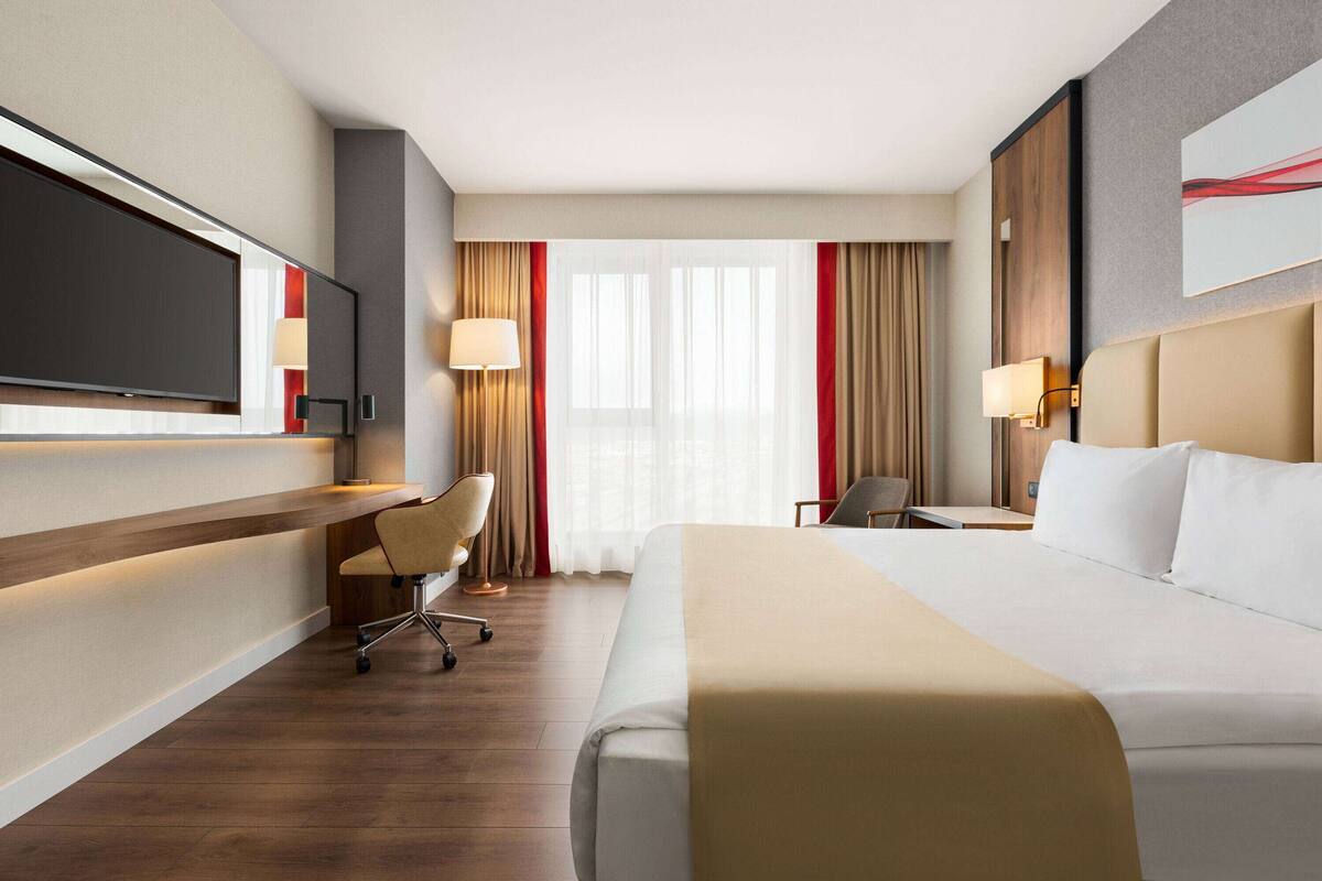 A hotel room with a large bed, a desk with a chair, a television, and a window with curtains.
