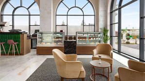 Een moderne hotel lobby met grote, gebogen ramen, een glazen vitrine met gebak, comfortabele zitplaatsen en een groene wand met barstoelen.
