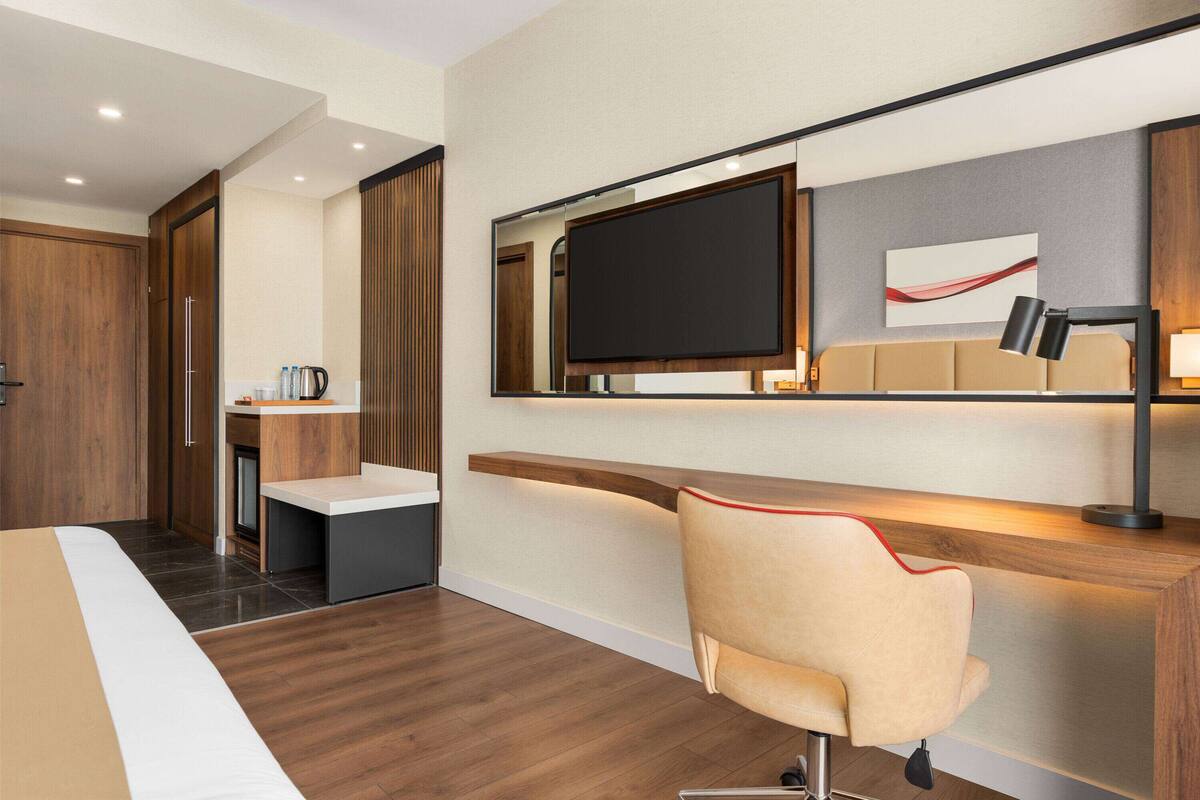 A modern hotel room with a wooden floor, a bed with white and beige bedding, a desk with a chair, a flat-screen TV mounted on the wall, and a coffee machine on a small table.