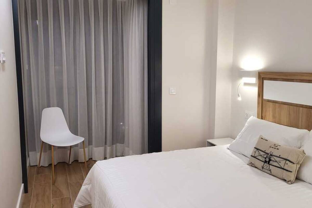 Una habitación de hotel con cama, una silla, una cortina y una lámpara fijada a la pared.