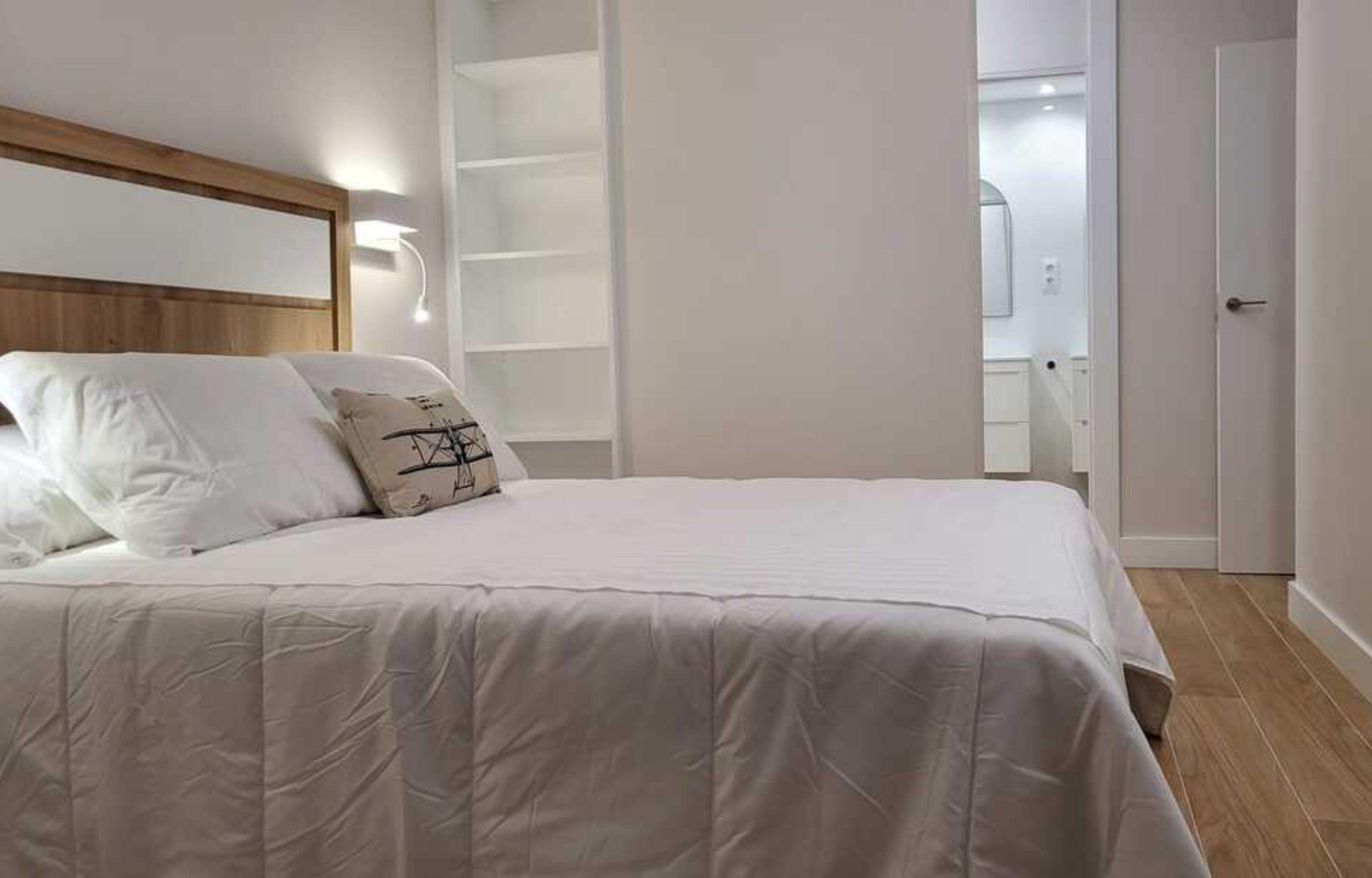 Un dormitorio con una cama grande, cabecera de madera, unidad estantería empotrada y una puerta que da al baño.